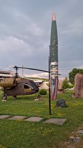 A museum park displays military aircraft and weapons. A helicopter is parked on the grass, with a missile standing vertically beside it. In the background, buildings and trees are visible, with more aircraft scattered throughout the park. The sky is cloudy.