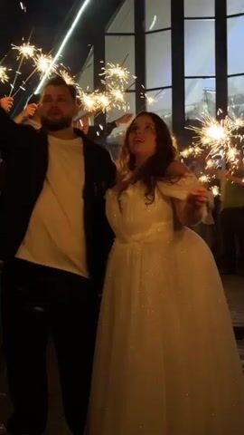 A man and a woman are standing close together outdoors at night, surrounded by people holding sparklers. The man is wearing a black suit jacket over a white shirt, and the woman is wearing a white wedding dress. They both appear to be looking up and to the right. The background shows a dark sky with some building lights. The sparklers are creating a bright, festive atmosphere.