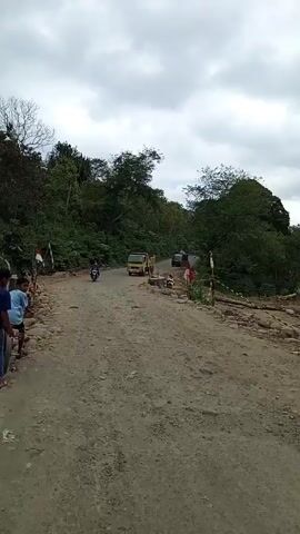 The video depicts a damaged, unpaved road with debris scattered around it. Two boys stand on the side of the road, one bent over a bicycle. A yellow truck and a motorcycle approach from the distance. Trees line both sides of the road, and the sky is overcast. People can be seen standing further up the road, near the truck. An explosion is briefly heard and a vibration detected.