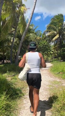 A person walks away from the camera on a narrow dirt path between tall grass and palm trees. The person is wearing a white tank top, black shorts, a baseball cap, and flip flops. They are carrying a cream-colored bag over their shoulder. The sky is blue with scattered white clouds. The scene appears to be a tropical area.