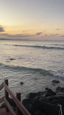 The video shows a view of the ocean at sunset from a slightly elevated perspective, likely a wooden walkway or viewing platform. In the foreground is a section of wooden railing, slightly angled to the left. Below the railing, large, dark rocks are visible, and small waves are washing onto them. 

The ocean occupies the majority of the frame, with several surfers visible in the distance. They are scattered across the water, some riding waves and others seemingly paddling or waiting. The waves are small and gentle, breaking softly.

The sky is a gradient of colors, with warm orange and yellow hues near the horizon blending into a lighter gray-blue towards the top of the frame. There are scattered clouds, and what appears to be a cityscape is faintly visible on the horizon, far in the distance.

The overall scene has a calm and peaceful atmosphere. The camera is mostly stationary, offering a steady view of the sunset and the surfers in the water.