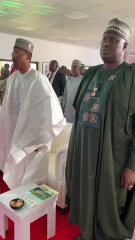 The video shows a group of men standing in what appears to be an indoor event space. Most of them are wearing traditional clothing. A red carpet covers the floor. Small white tables are placed near each individual, each holding a book and a dish of small snacks. A few people in the background wear suits. The overall impression is of a formal or official gathering.