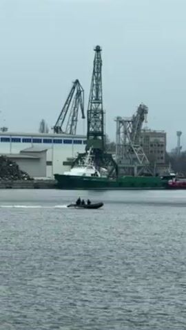 In a medium shot taken from a stationary position, a black inflatable boat with a small number of people aboard navigates across a body of water in front of a harbor or port. The water's surface is rippled from the boat's movement. In the background, a series of large, green industrial cranes are visible against an overcast sky. The foreground consists of the water and the boat, while the background contains the cranes, various buildings, and what appears to be industrial storage. The overall atmosphere of the scene is somewhat bleak due to the muted colors and the overcast conditions. The boat is the main moving element, creating a sense of activity against the static industrial backdrop.