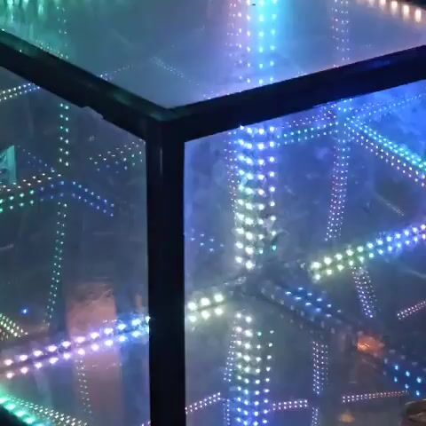 The video shows a close-up of an infinity mirror cube. The cube is lit with multicolored LEDs that create an illusion of infinite depth. The lights are cycling through various colors, including blue, green, and orange. The cube is made of glass and has a black frame. The background is blurred and dark.