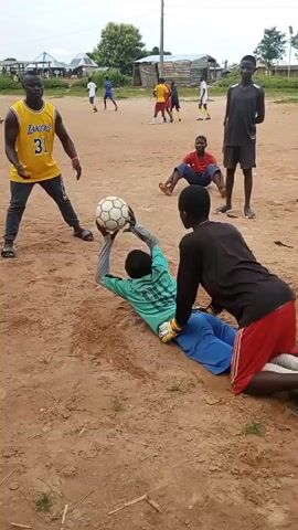 The video shows a group of young men playing soccer in an outdoor dirt field. In the foreground, a young man in a yellow Lakers jersey with the number 31 on it is holding a soccer ball. He is facing another young man who is on his hands and knees, wearing a green shirt and blue shorts, and appears to be acting as a goalkeeper. A third young man in a black shirt and red shorts is kneeling behind the goalkeeper. In the background, several other young men are scattered across the field, some appearing to be playing soccer or watching. The environment is a dusty, unpaved area with some buildings and trees visible in the distance. The sky is overcast.