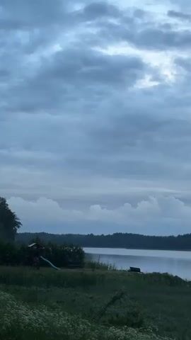 The video shows a serene lake scene with a cloudy sky. In the foreground, there is lush green grass and some white wildflowers. To the left, a small playground structure with a blue slide is visible, along with a wooden bench. The lake stretches out into the distance, with a dense, dark forest lining the opposite shore. The water is calm, reflecting the muted colors of the sky. The overall impression is one of tranquility and nature.
