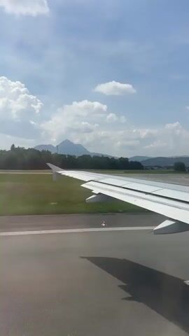 The video captures a view from an airplane window during takeoff at Salzburg Airport, Austria. The primary subject is the airplane's wing, which dominates the foreground. Beyond the wing, the runway stretches out, marked with yellow lines and white stripes. A smaller aircraft is visible on a parallel taxiway in the distance. The background features a lush green landscape with trees and scattered buildings, leading up to rolling hills and a prominent mountain peak under a bright, partly cloudy sky. The time of day appears to be late morning or early afternoon, given the clear and sunny weather. The scene evokes a sense of anticipation and the beginning of a journey. There is no visible text in the media file.