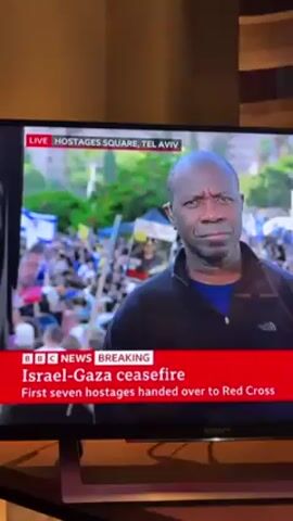 A television screen displays BBC News coverage of the Israel-Gaza ceasefire. The breaking news banner at the bottom of the screen reads, "Israel-Gaza ceasefire. First seven hostages handed over to Red Cross." The live location is identified as Hostages Square, Tel Aviv. A man with dark skin is featured in the foreground, seemingly reporting live from the scene. Behind him, a crowd is visible, with many holding what appear to be flags. The television is a Sony brand, and is resting on a wooden table.
