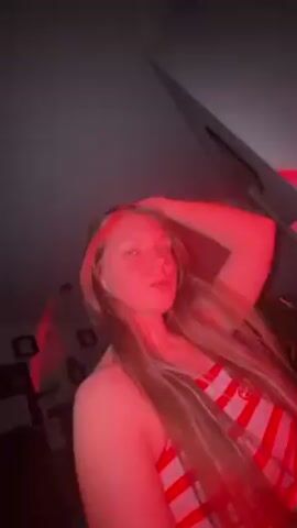 A woman with long, light brown hair is illuminated by a red light. She wears a red and white striped top. She looks at the camera and touches her face. The background is dark, with a ceiling fan visible in the corner. There are framed pictures on the wall behind her.