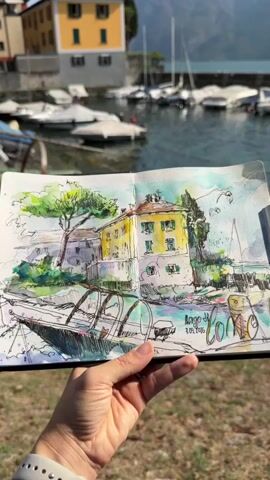A hand with a white smartwatch holds open a sketchbook with a watercolor and ink sketch of a yellow building with white trim, surrounded by trees, and a boat in the foreground. In the background, there are several boats covered with white tarps on a body of water, with a mountain range visible behind them. The scene is outdoors, with sunlight casting shadows. The sketch includes handwritten text that appears to read "Lago di Como" and a date.