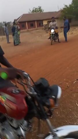 The video appears to be shot from the perspective of a person riding a motorcycle on a dirt road in what looks like a rural area. Several people are standing on the side of the road, and another person is sitting on a motorcycle. The person filming the video then starts riding their motorcycle down the road. There are buildings in the background and some debris on the ground. The video is shaky due to the movement of the motorcycle.