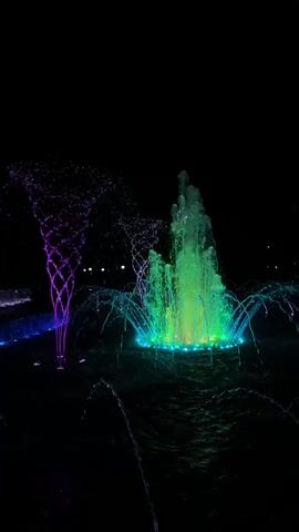 The video captures a night scene of a fountain with colorful lights. The fountain in the foreground emits water in arcs and streams, illuminated by green and blue lights, creating a vibrant glow. To the left, another fountain sprays water in a swirling pattern, also lit with blue and purple hues. The background is dark, suggesting it's nighttime, with a few distant lights visible, possibly from streetlights or buildings. The water's surface reflects the lights, adding to the dynamic visual. There are no people or animals visible in the frame. The focus is entirely on the illuminated water features.