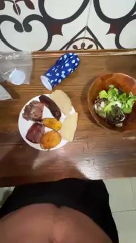 Here is a description of the video.
The video begins with a shot of a table featuring a plate with meat, sausage, fried plantains, and what appears to be arepas. Next to it is a bowl of salad, a blue patterned pouch, and a plastic bag. The camera moves upward to show a man with dark hair, a mustache, and a shirtless torso smiling at the camera. The background is blurry but shows lights and a ceiling.