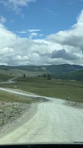 The video shows a view from inside a vehicle driving on a gravel road. The road curves through rolling green hills under a cloudy blue sky. In the distance, there are more hills and mountains, some covered in trees. The vegetation appears to be grass and sparse trees. The sky has dramatic white clouds scattered across the blue. The perspective is from the driver's seat looking forward and slightly to the right.