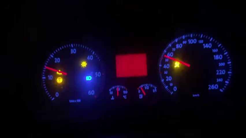 The video shows a close-up of a car's dashboard at night. The dashboard lights are illuminated, with a blue glow around the gauges. The speedometer needle is pointing to approximately 40 km/h, and the tachometer needle is at around 2000 RPM. Several warning lights are also lit: a yellow symbol resembling a car with a lock inside (likely an immobilizer or security light), a yellow symbol of a light bulb with an exclamation mark (likely a bulb failure warning), and a blue symbol of a headlight (likely the low beam headlight is on). A red warning light at the top center, shaped like a thermometer, is also illuminated, indicating a potential engine overheating issue. The central digital display is blank and red. The background is dark, suggesting the car is in motion on a road at night.