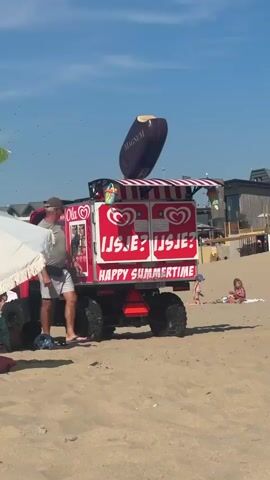 A red ice cream truck with "Ola" and "Happy Summertime" written on it is parked on a sandy beach. A large inflatable Magnum ice cream bar is displayed on top of the truck. A man in a grey t-shirt and shorts stands near the truck, looking at it. A young boy in patterned shorts walks towards the truck. In the background, children are playing on the sand. The sky is blue with some clouds.