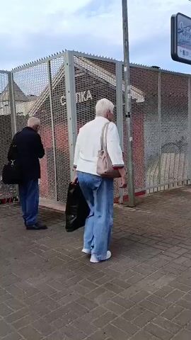 The video shows a street scene with two older adults walking away from the camera. The person on the left is wearing a dark jacket and jeans, carrying a dark bag over their left shoulder. The person on the right is wearing a white sweater, jeans, and white shoes, carrying a light-colored bag over their right shoulder and a black bag in their right hand. They are walking past a metal fence. Behind the fence, there is a red structure with the word "СПАСКА" written on it in white letters. A man in a dark blue polo shirt, jeans, and headphones around his neck stands facing away from the camera, looking to the right, with a backpack on. The ground is paved with brick-like tiles. In the background, there are buildings and a blue sky with clouds. A sign with Cyrillic writing is visible in the upper right corner.