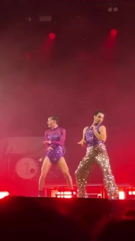 Two men are performing on a stage bathed in red light. The man on the left is wearing a shiny purple bodysuit with long sleeves, while the man on the right wears a matching purple vest and gold sequined pants. The man on the right is singing into a microphone, making gestures as he sings. The other man is dancing in sync, making similar movements. A drum kit is visible behind them on the left side of the stage. The foreground shows the audience, partially obscuring the lower part of the stage. There are stage lights above, contributing to the red glow of the scene.