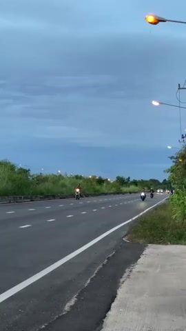 A vertical video captures a highway scene at dusk or dawn. The sky is a mix of blue and grey clouds, with streetlights starting to illuminate. The road is paved with white lines. Several motorcycles are visible on the road. In the foreground, a person rides a white scooter. Further down the road, two more motorcycles are present. The sides of the road are lined with lush green grass and trees. The camera pans slightly upwards, showcasing the vast sky and the emerging lights of the street lamps.