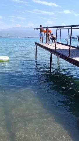Two children are on a wooden pier over a body of water. One child, wearing arm floaties, is standing with their back to the camera. The other child, facing the camera, is standing near the edge of the pier with their arms outstretched. The second child jumps into the water, creating a splash. The water is clear, revealing the sandy bottom. Mountains are visible in the distance. The sky is blue with some clouds.