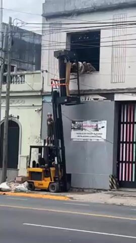 The video shows a construction site with a yellow Clark forklift carrying a platform with a man standing on it, operating the lift. At the top of the lift, two men are working on a second-story window. One man is inside the building, reaching out to grab materials, while the other is on the platform, handing them over. The building is old and has a white exterior. The scene is outdoors, and a street runs along in front of the building, with some traffic passing by. A large sign is next to the lift, advertising "Maderas el Colorado." There are power lines and other buildings in the background.