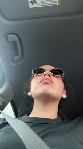 The video shows a woman seated in a car, wearing sunglasses, a black t-shirt, and pearl earrings. She is wearing her seatbelt. The camera angle is low, looking up at her. Car interior elements such as the ceiling, handle, and visor warning label are visible. The woman appears to be looking upwards. The sound of music is audible.