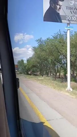 The video shows a view from inside a vehicle looking out the passenger side window. A paved road with a yellow dividing line runs alongside the vehicle. On the right side of the road, there are trees and dry vegetation. In the distance, there is a billboard with text and an image that appears to be an advertisement. The sky is blue with some white clouds. The video appears to have been filmed while the vehicle was in motion.
