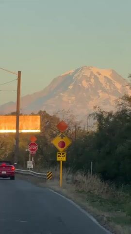 A red car is driving down a road in the foreground with a mountain in the background. There are several traffic signs including stop signs and a speed limit sign for 25 MPH. The sun is setting and casting a warm glow on the mountain and the sky is a clear, pale orange. The mountain is snow-capped and appears to be a dormant volcano. The trees are bare and the grass is dry, indicating it is likely autumn or winter. The overall scene is peaceful and serene.