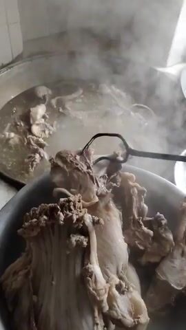 A close-up shot captures large pieces of meat with bones, possibly from a whole animal, submerged in a steaming pot of broth. A metal utensil with two prongs is shown stirring the contents of a larger pot in the background, where more meat and broth are visible. Steam rises from the pots, indicating the food is being cooked or has just been cooked. The scene appears to be in a kitchen or food preparation area.