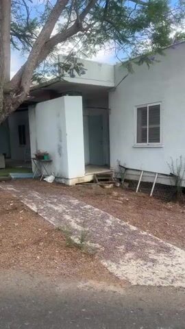 The media file shows a first-person perspective video of a person walking along a path and then a road in an outdoor setting. The scene transitions between different views, suggesting movement and exploration.

Initially, the view focuses on a simple, white, rectangular building with a small window and a door. The area around the building is covered with gravel and some patchy vegetation. In the foreground, a path is visible, partially covered with fallen purple flowers, possibly from a jacaranda tree, which is also visible in the frame.

The perspective then shifts to a wider view, revealing more of the surroundings. Lush green trees, including a large one with vibrant green foliage, dominate the landscape. A dark-colored car is parked under the tree, and a white plastic chair sits nearby. A street lamp is also visible. The ground is a mix of gravel and dirt.

Further along, the scene opens up to a road curving gently. More greenery and trees line the sides of the road, with a glimpse of distant hills or landscape in the background. The sky is blue with scattered white clouds, indicating it's daytime, likely morning or afternoon. The overall atmosphere appears calm and peaceful.

The final segment of the video shows the perspective of someone walking on an asphalt road. A calico cat with a black-tipped tail walks ahead on the road, appearing to lead the way. The road is bordered by vegetation and a paved area on the side.

There is no visible text in the media file. The setting appears to be a semi-rural or suburban area, possibly a residential compound or a public space with natural elements. The presence of blooming flowers and the greenery suggests a pleasant weather condition.