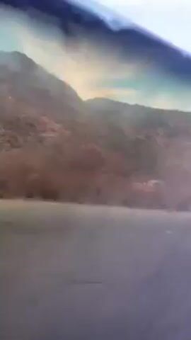 The video shows a view from inside a moving vehicle, possibly a car, looking out towards a body of water and a mountain range. The mountains are rocky and barren with sparse vegetation. The water appears calm, reflecting the sky and surrounding landscape. The sky is pale blue with some clouds. There are occasional reflections on the window of the vehicle.