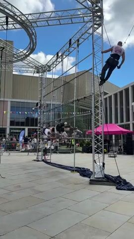 A group of performers executes aerial maneuvers on a climbing apparatus in an outdoor public square. The apparatus consists of a tall metal frame with a circular structure on top, and multiple net sections stretched between the frame's vertical supports. Several people are performing simultaneously; one person hangs by their leg, another is mid-air in a backflip, and another is suspended at the top of the frame. Below, a black tarp is spread on the ground. A crowd of onlookers is visible behind a safety barrier. In the background, there is a building with banners and a Starbucks sign. A pink pop-up tent sits beside the apparatus. The sky is blue with scattered clouds.