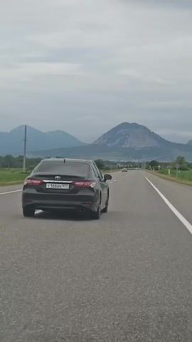 A black car is driving on a road. In the background, there are mountains. The sky is cloudy. The car has a license plate that is visible. Another white car passes the black car.