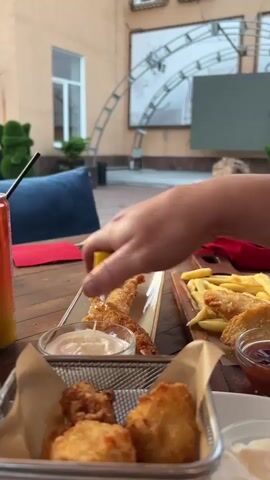The scene shows a person squeezing lemon juice onto breaded chicken strips on a restaurant table. The table is set with multiple dishes including a metal basket with chicken nuggets and ramekins of sauce. A can of soda with a straw is visible. In the background, a child is briefly seen running in an outdoor patio setting.