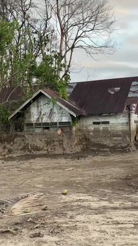 The video shows a scene of destruction, likely caused by a natural disaster. The ground is covered in mud and debris, with several houses partially buried or damaged. The houses appear to be made of wood and tin, and some have their roofs caved in or tilted. Trees are visible in the background, some of which are bare and appear dead. The sky is cloudy with some patches of light, suggesting it might be early morning or late afternoon. There are no people visible in the video. The overall impression is one of devastation and loss.