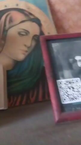 A still image shows a painting of a religious figure, possibly the Virgin Mary, with a halo. To the right of the painting is a framed picture, which appears to be a black and white photograph. A QR code is visible on the framed photograph. To the left of the painting is a light-colored object that could be a book or a stand. The objects are placed on a dark surface, possibly a shelf or a table. The background is a textured surface, perhaps a wall.