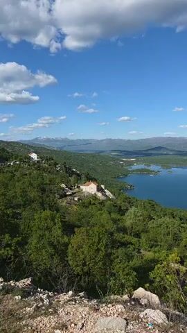 The video shows a wide, scenic view of a lake with several small, green islands scattered throughout. The water appears calm and a deep blue color. The foreground is filled with lush green trees and shrubs, providing a natural frame for the lake. In the distance, mountains can be seen, some with visible snow cover. The sky is a clear, bright blue with a few scattered white clouds. A couple of buildings are visible nestled amongst the trees on the hillside in the middle ground. The camera pans slowly across the landscape, revealing more of the lake and the surrounding nature. The environment appears peaceful and untouched.