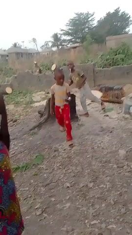 The video shows a dirt-filled yard.  A young boy in a t-shirt and red pants is dancing in the foreground.  Another boy wearing a t-shirt and light-colored pants, stands behind him with something in his mouth. Then, the second boy approaches the camera with a cigarette-like object in his mouth.  The camera follows him as he walks toward a building. In the background are tree stumps, logs, and a fence.