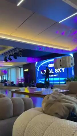 The video shows the interior of a restaurant or lounge-like venue with modern lighting and decor. A stage area is visible in the background, featuring a large screen displaying the name "Royal Sky Restaurant" and a person performing on what looks like a keyboard. The venue has various seating areas with round, cushioned furniture. People are scattered around the room, seated at tables or walking around. There are decorative plants, columns, and linear lighting fixtures on the ceiling which change color from purple to green. The video appears to be taken from a seated position, with a focus on the overall ambiance and the presence of people within the space.