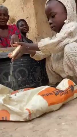 A young child, dressed in a hooded sweater, sits on the ground, carefully pouring rice into a large white bag with orange lettering and elephant images. The child is surrounded by adults, including an older woman in a red and pink top who watches the child intently. The scene takes place outdoors, possibly in a courtyard or similar open space, with textured walls in the background. The atmosphere appears communal, with the activity of sorting rice taking place in a shared space.