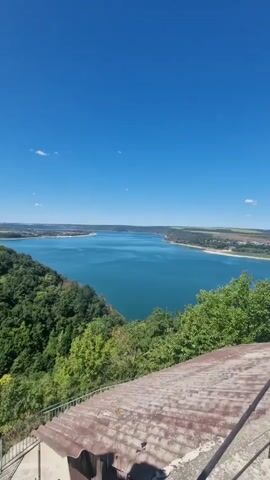 This is a vertical, wide shot from a high vantage point overlooking a vast, blue body of water, likely a lake or reservoir, under a clear blue sky. The water stretches into the distance, meeting a flat, green, and brown horizon. On the left, a steep, densely wooded hillside covered in lush green trees slopes down towards the water. In the foreground, the top of a building with a rusty, corrugated metal roof is visible, along with parts of its concrete structure and a metal railing. The sunlight appears bright, indicating it is daytime. There are no people visible in the video.