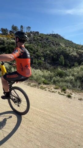 A man wearing a cycling helmet, an orange and black cycling shirt with “BILABIKERS” printed on the back, and black cycling shorts rides a yellow and black mountain bike down a dirt path. He's performing a wheelie, lifting the front wheel off the ground. The man looks to his right, then faces the camera. He sticks his tongue out and flashes a rock-on sign with his hand. The sun is shining in a blue sky and there is dry vegetation and large rocks on the hillside behind the path. Another cyclist passes behind the first man on the right.
