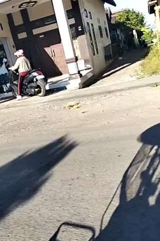 The video shows a street scene with several people and vehicles. In the foreground, a motorcycle is parked or being ridden, casting a shadow on the asphalt road. Further down the street, two individuals are standing near a building that appears to be a shop or a small business, with shelves visible through a window. One person is wearing a white uniform, possibly a school uniform or medical attire. Another person is standing slightly behind them. A second motorcycle is also present, with a person wearing a red jacket and a white head covering riding it. The environment appears to be a residential or commercial area, with buildings and trees visible. The lighting suggests it is daytime, with bright sunlight.