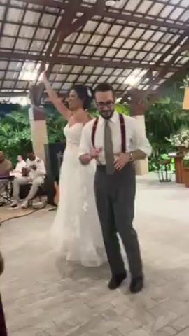 The video shows a wedding reception held under a covered patio. A bride in a white strapless gown and groom wearing gray trousers, a white shirt with suspenders, and glasses, are standing on a stone-tiled floor, dancing to music performed by a live band. The groom has a tie. Two women in dresses, one maroon and one light green, are also dancing. People are clapping and cheering the bride and groom. The patio has exposed wooden beams and is surrounded by green foliage. The band is seated in the background. The lighting is relatively dim.