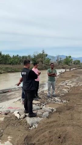 Four people are standing on a dirt path next to a muddy river. The ground is covered in dirt and rocks. In the background, there are trees, mountains, and construction equipment, including an excavator and a yellow truck. The sky is cloudy.