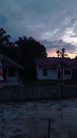 The video shows a twilight scene with two houses and palm trees silhouetted against a cloudy sky. The sky displays hues of purple, pink, and gray, indicating either sunrise or sunset. Both houses have red roofs and white walls. The house on the left is partially visible, while the house on the right is more prominent and features decorative trim along the roofline and small flags attached to poles in front of it. A low concrete fence separates the properties from the foreground, which appears to be a wet, paved area with reflections. The overall atmosphere is calm and serene.