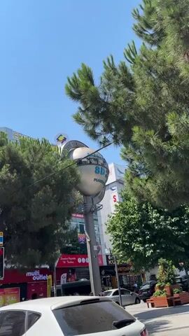 The video shows a large, silver, spherical monument with text and images on it, standing on a pole. It is surrounded by trees and buildings. The monument appears to be in a city, with traffic and signs visible. The text on the monument appears to be in Turkish. The sky is blue and the sun is shining.
