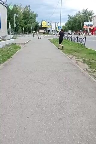A person is running on a sidewalk. There are trees and buildings in the background. There is also a fence and grass. The person is wearing a black t-shirt and black shorts. The person is running away from the camera.