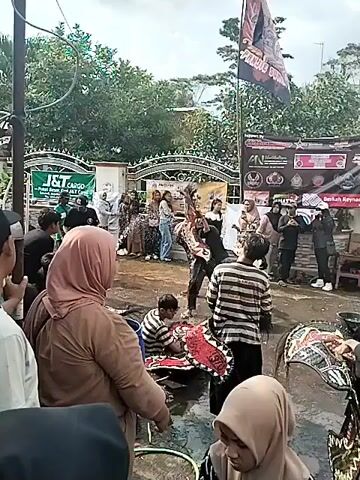 Here's a description of the video:

The video shows a street scene with a group of people gathered around several individuals who appear to be performers or participants in a cultural event. Several people are on the ground, interacting with what looks like a red and black ornate object, possibly part of a costume or prop. Water is being splashed around. Other people stand around watching.

In the background, there's a building with a J&T Cargo sign, a decorative gate, and a banner hanging from a pole. The ground appears wet, possibly from the water being splashed or from recent rain. People are dressed in casual clothing and some women are wearing hijabs. The overall atmosphere seems festive and active.