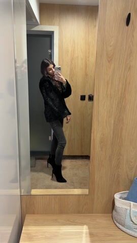 A young woman stands in front of a mirror, taking a selfie. She is wearing a black, furry jacket, a black turtleneck sweater, grey skinny jeans, and black heeled boots. The mirror is framed with wood and is mounted on a light wooden dresser. To the right of the mirror is a wooden closet door, and to the left is a grey door. A white basket containing a blue item is sitting on the dresser to the right of the mirror.  The woman holds a smartphone in her hand. She is facing the mirror, looking at her reflection.
